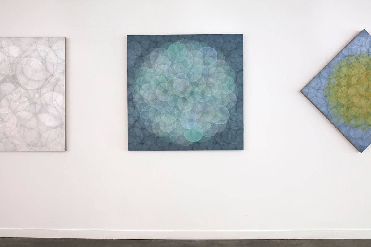 A blue, teal, light lavender and white abstract geometric painting hung on a white wall in strong lighting.