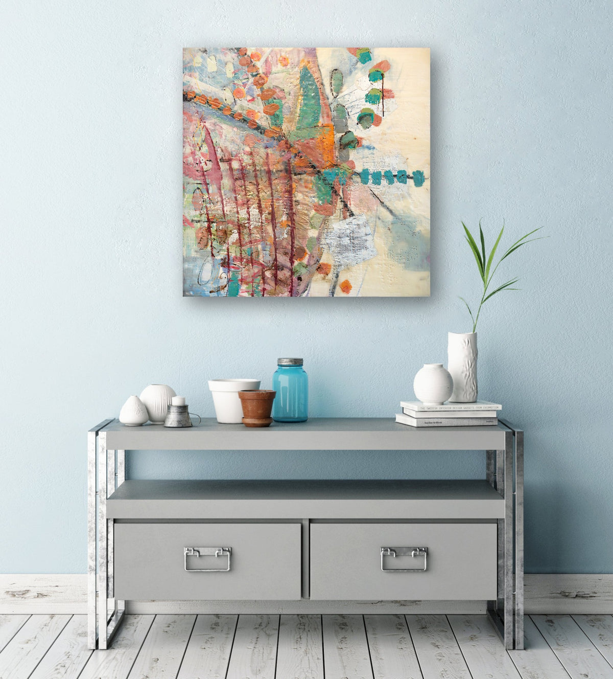 A multicolored abstract print hanging on a light blue wall above a grey accent table, pots, vases, and stacked books. 