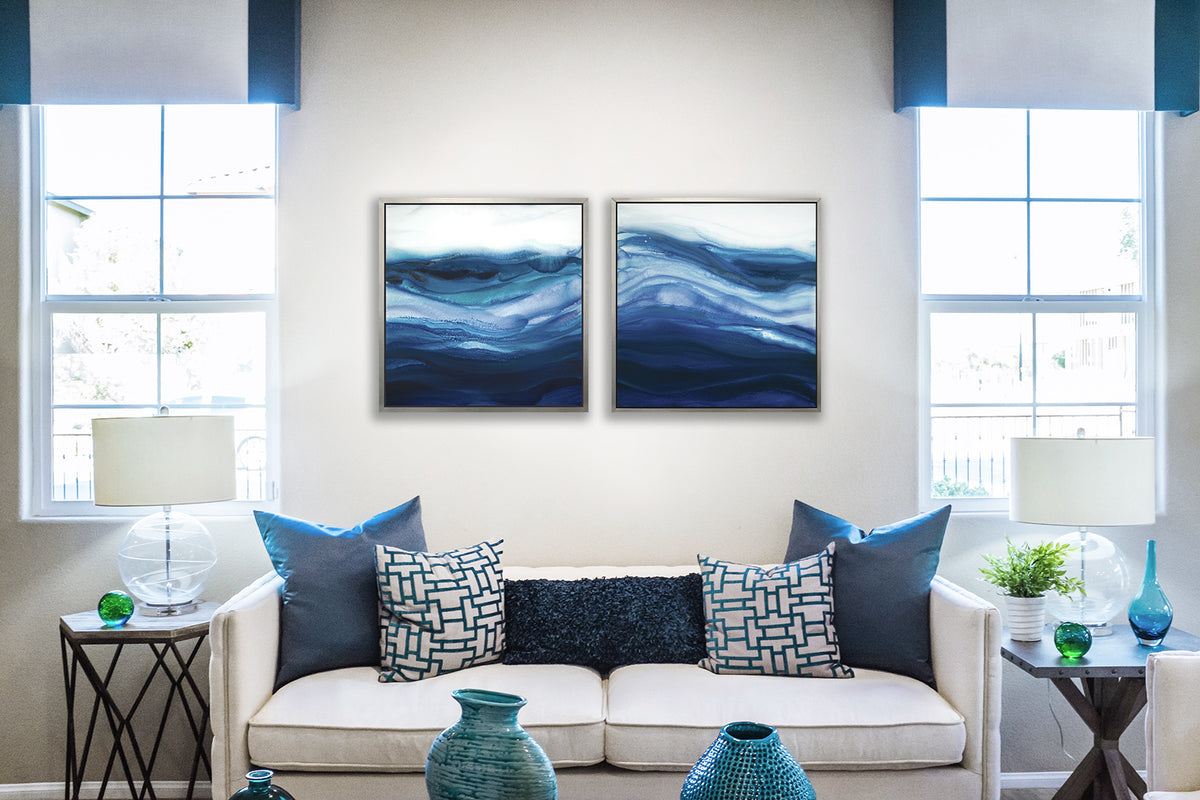 Two blue, teal and white abstract prints in silver floater frames hang side by side on a white wall above a white sofa with blue pillows and side tables with table lamps.