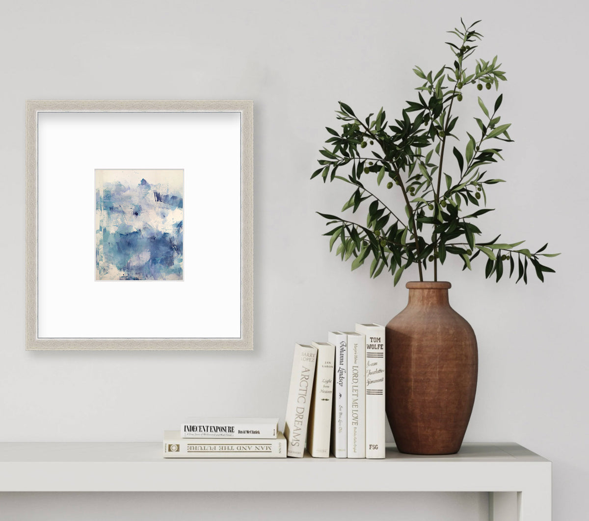 A framed abstract paper print hangs on a white wall above a console table with books and leafy bouquet in a brown vase. 