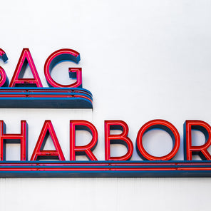A photograph of a blue and red neon sign that reads "Sag Harbor" on the side of a white wall. 