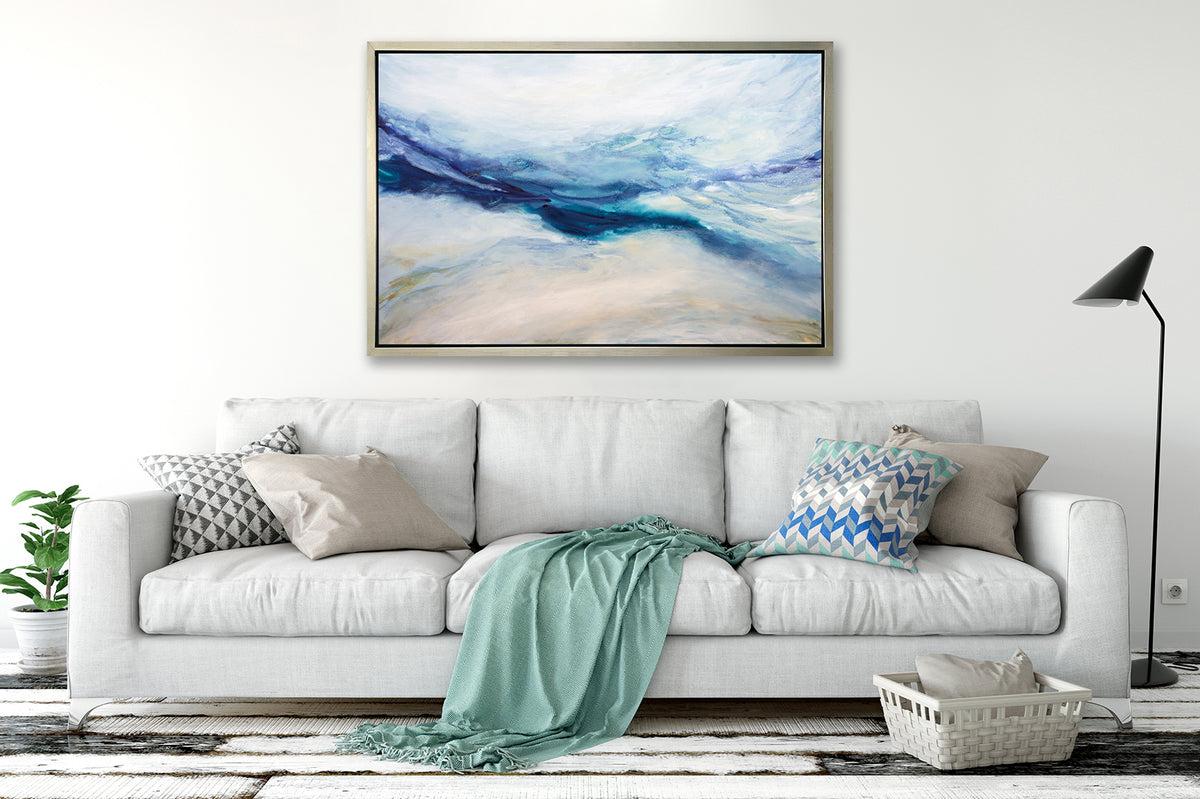 A white, blue, purple and beige abstract print in a silver floater frame hangs on a white wall above a white sofa with printed pillows, a light green throw blanket and a tall black floor lamp.