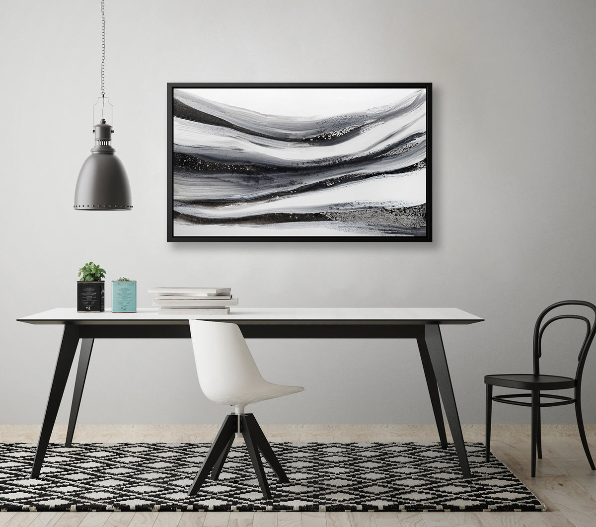 A black, white and grey abstract print in a black floater frame hangs on a white wall above a white table with a stack of books, two potted succulents, a white chair and a black chair.