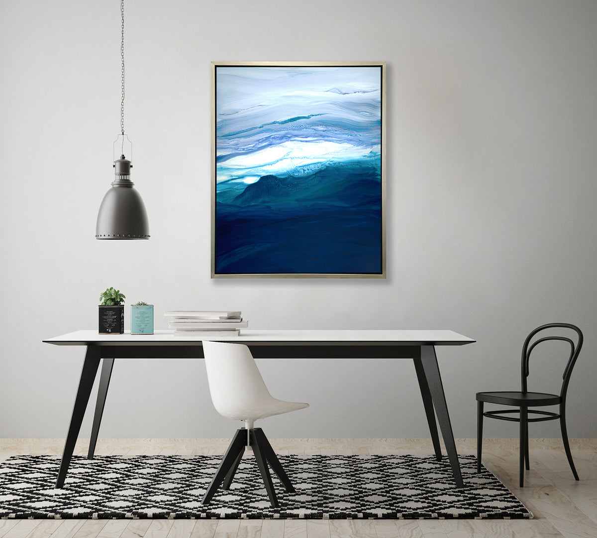 A blue, teal, lavender and white abstract print in a silver floater frame hangs on a white wall above a white table with a stack of books, two potted succulents, a white chair and black chair.