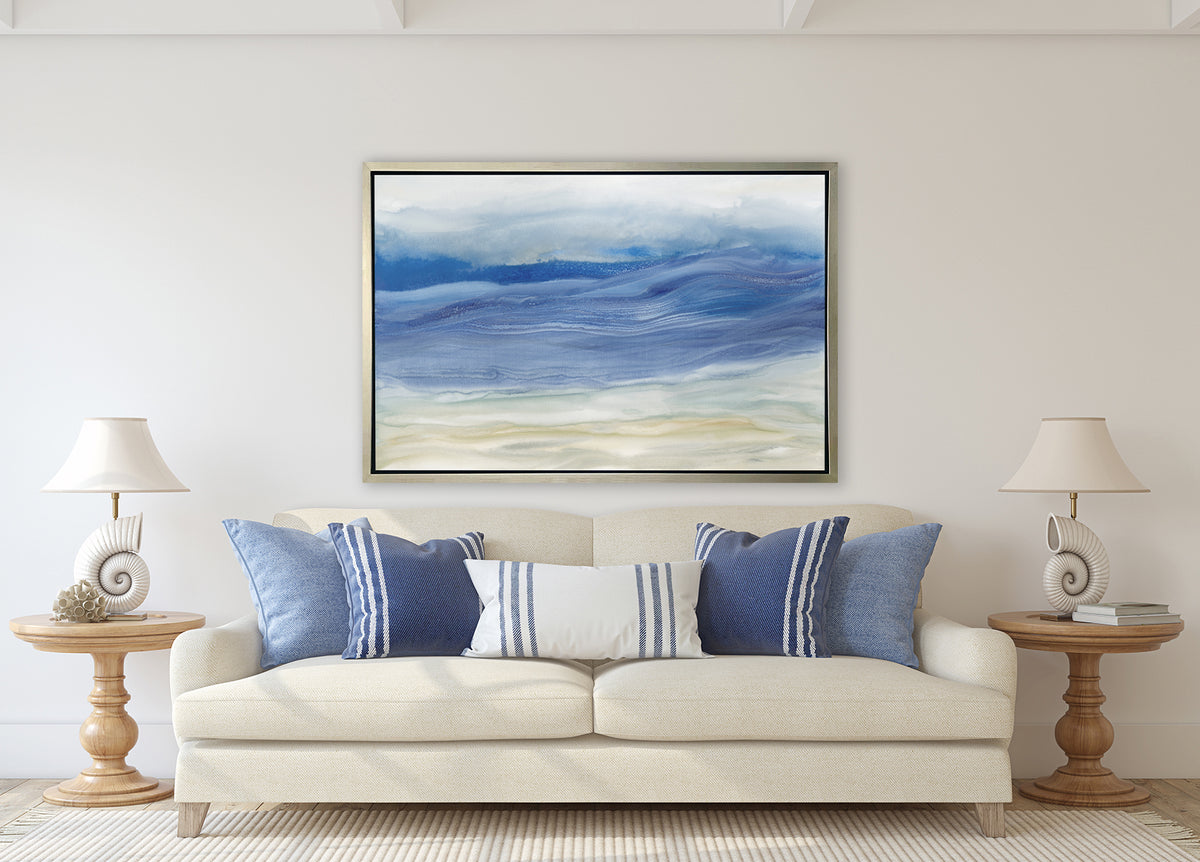 A blue, white and beige abstract print in a silver floater frame hangs on a white wall above a cream linen sofa with blue and white pillows and two matching wood pedestal side tables with matching white seashell table lamps.