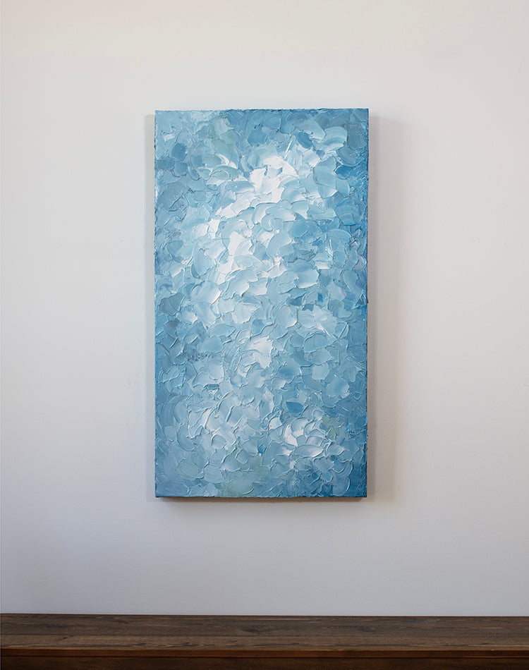A blue and white abstract painting by Teodora Guererra hangs vertically on a white wall over a bench.