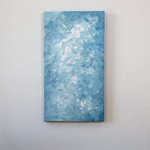 A blue and white abstract painting by Teodora Guererra hangs vertically on a white wall over a bench.