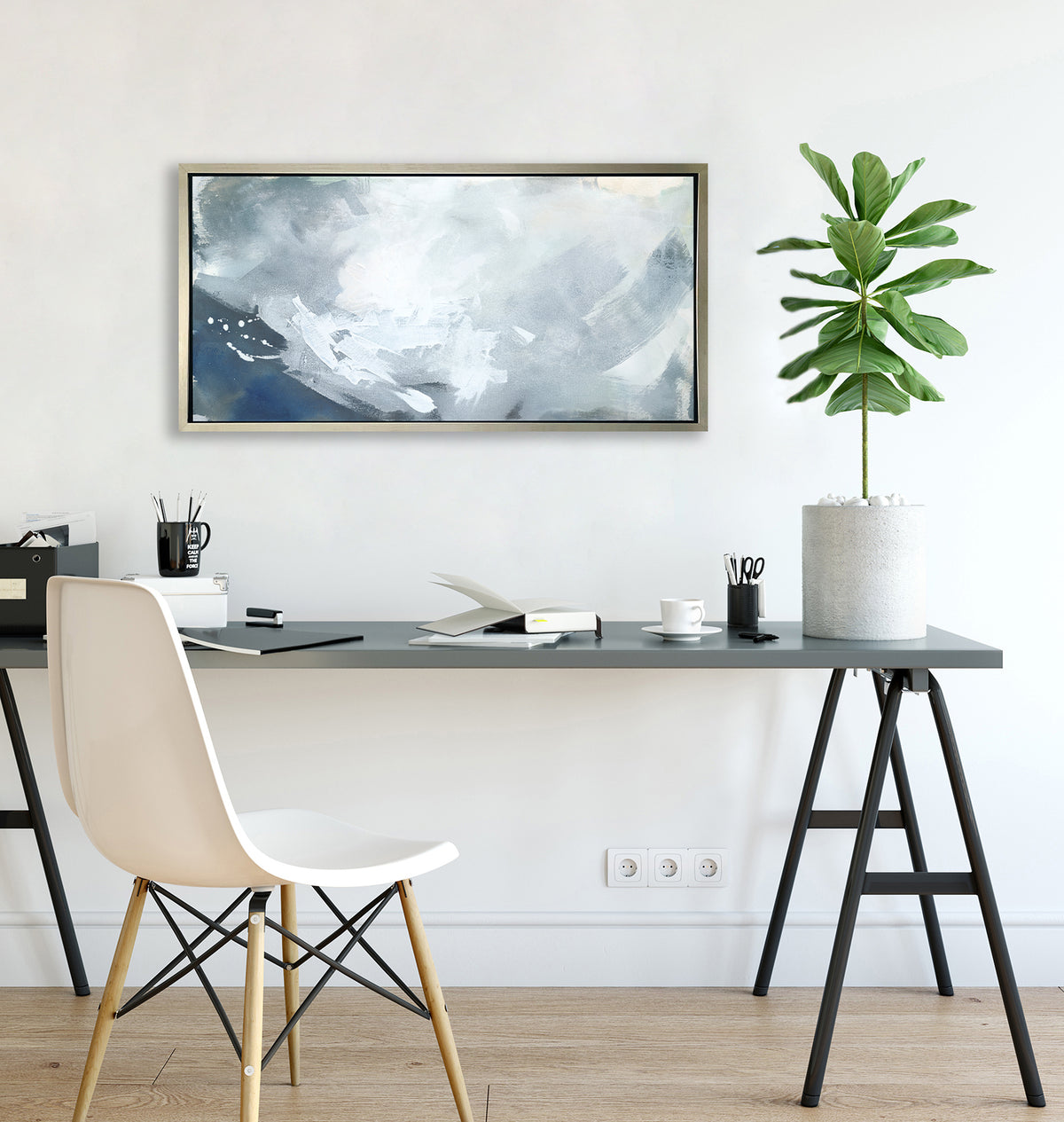 A blue, grey and white abstract print in a silver floater frame hangs on a white wall above a grey desk with open notebooks, a black mug with pencils and a potted plant.