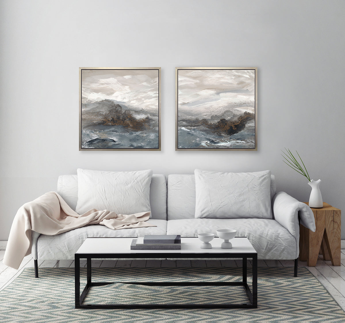 Two beige, white, brown and grey abstract prints in silver floater frames hang side by side on a white wall above a white sofa with pillows, a wood side table with a white vase and a white rectangular coffee table.