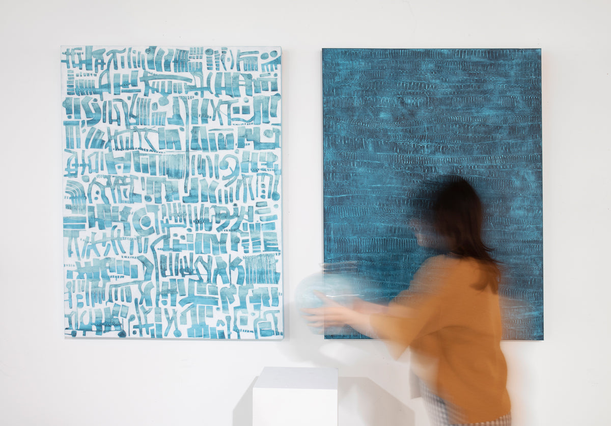 Two blue and white coastal abstract paintings hang on a white wall behind a woman in an orange top ads she places a sculpture on a pedestal in front of the paintings. 