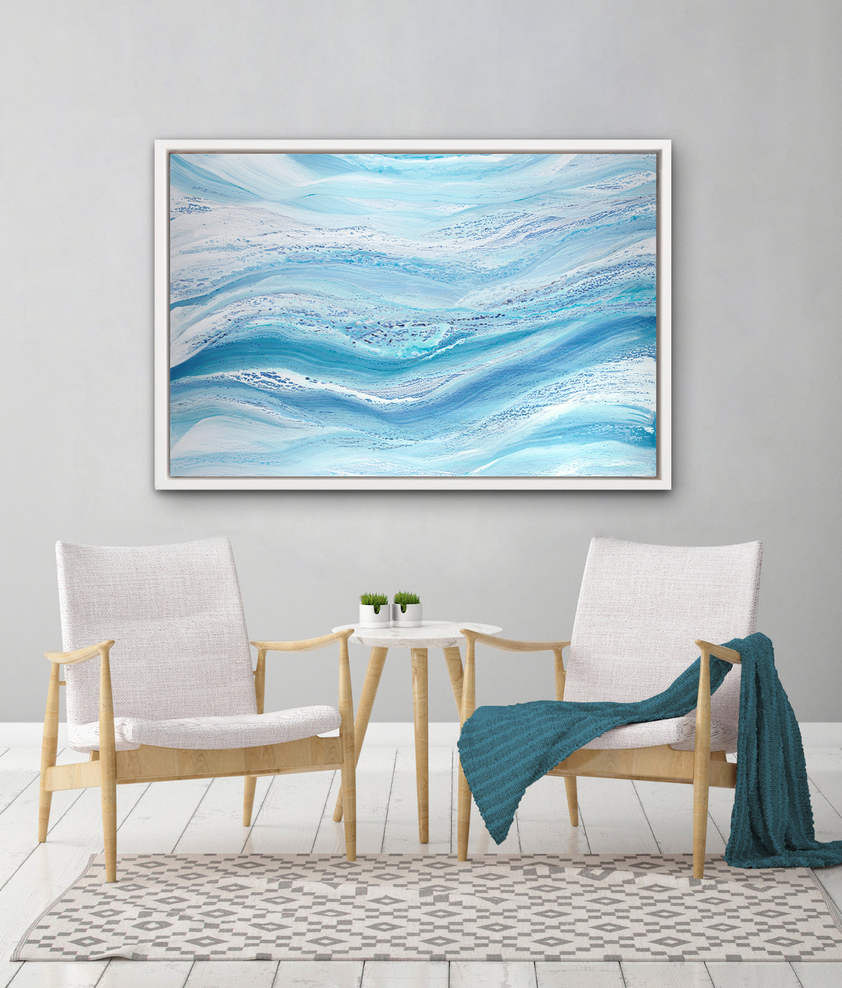 A blue, teal and white abstract print in a white floater frame hangs on a white wall above two white and light wood chairs with a dark teal throw blanket on one and a small white marble side table inbetween.