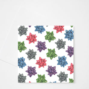 A white greeting card with red, green, blue, and silver bows displayed above a white envelope on a white background. 