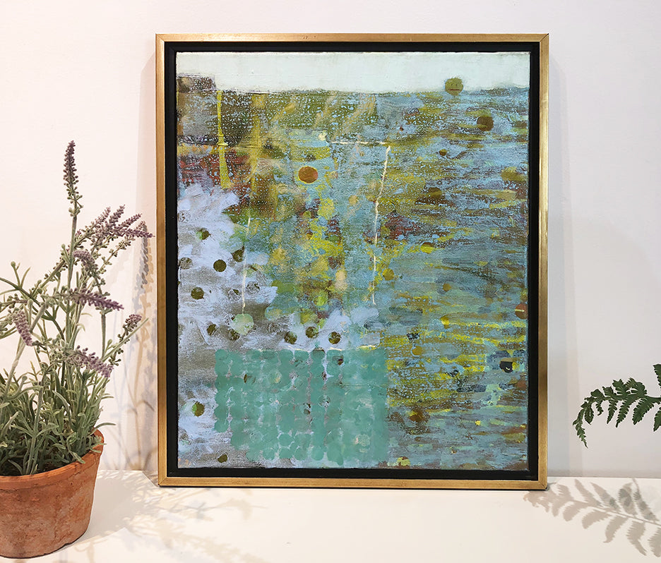 A green, blue and dark yellow abstract painting in a gold frame sits on a white surface next to a small potted plant.