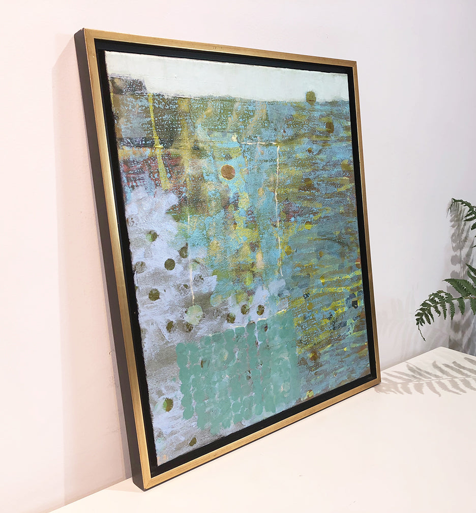 A left side-angle view of a blue, green and dark yellow abstract painting by Christine Averill-Green in a gold frame sits on a white surface.