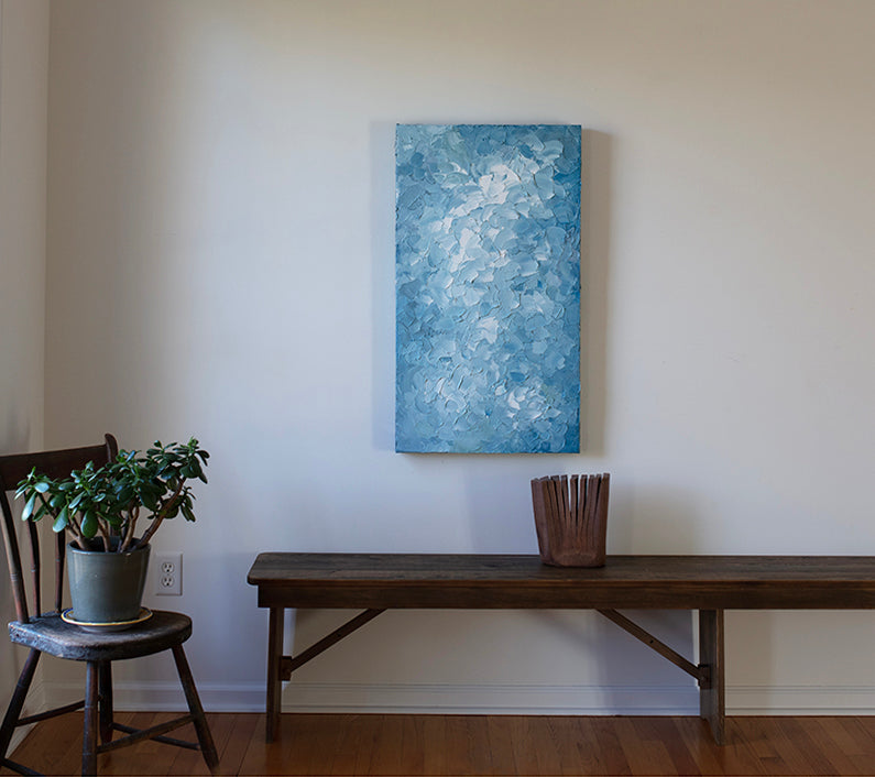 A blue and white textured abstract painting by Teodora Guererra hangs vertically on a white wall above a dark wood bench with a sculpture on the bench.  A chair is to the left with a plant on it.