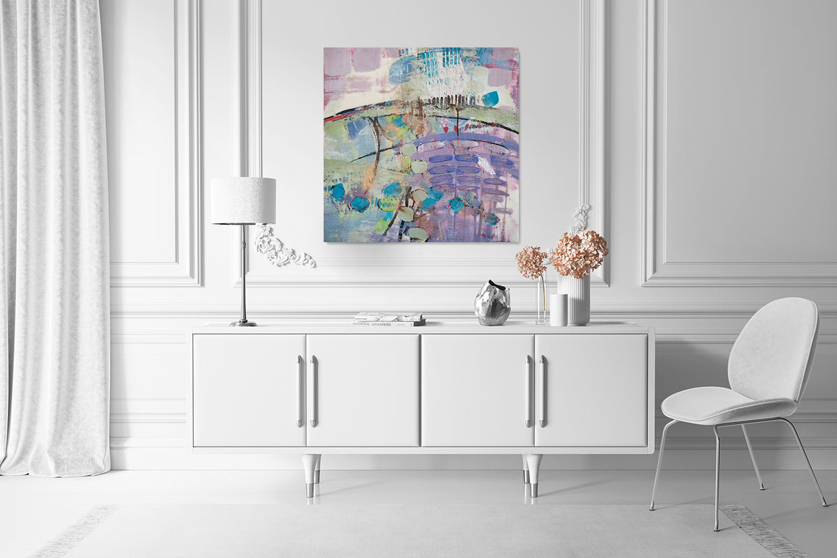 A purple, green, and blue abstract print by Linda Bigness hangs on white wall above a clean white console table, a lamp and decor accessories, and a white chair. 