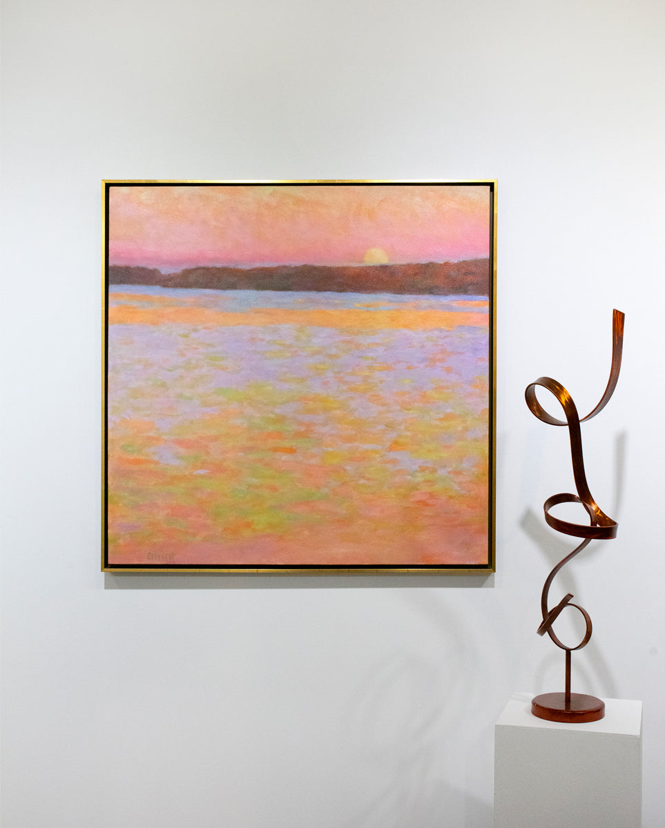 Framed painting of a sunset over water with a colorful sky hanging on a white wall with a sculpture to the side.
