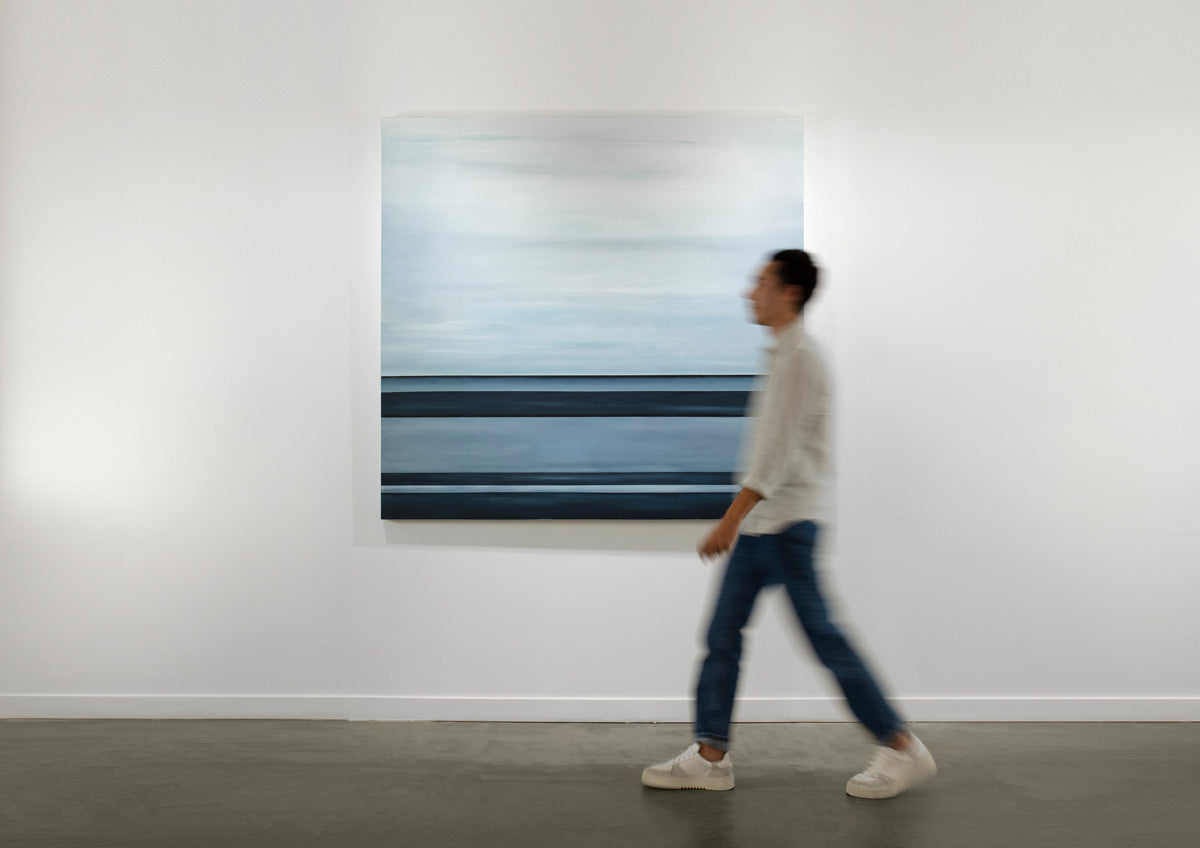 A tonal blue linear abstract painting by Tony Iadicicco hangs on a white wall as a person walks by.