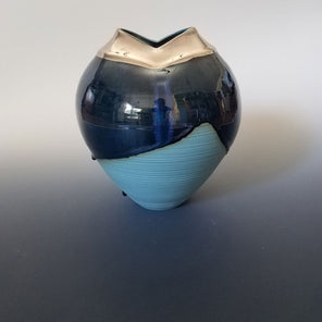 A light and dark blue sculptural vase with a 24-karat gold opening at the top sits on a clean grey surface. 